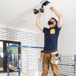 Electrician Builder at work, installation of lamps at height. Professional in overalls with a drill on the background of the repair site.